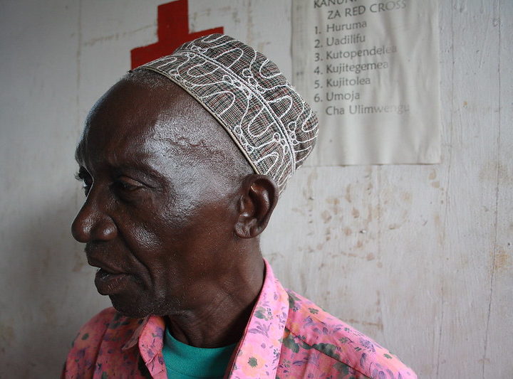 A man in a brightly coloured shirt and hat stands against a wall with a poster with the Red Cross logo