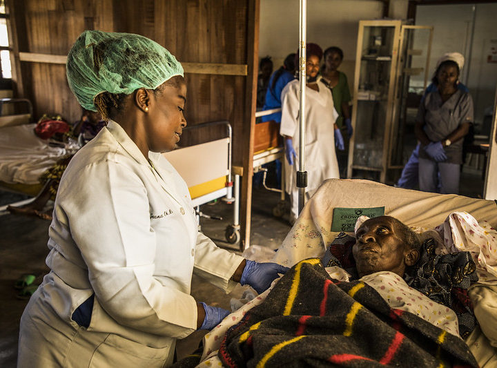 A health worker with a white coat and hair net touches a patient lying on a bed