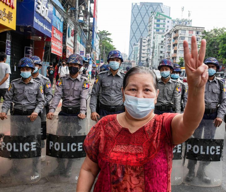 A woman in a red shirt and face mask makes a three-fingered salute in front of police officers during a demonstration.