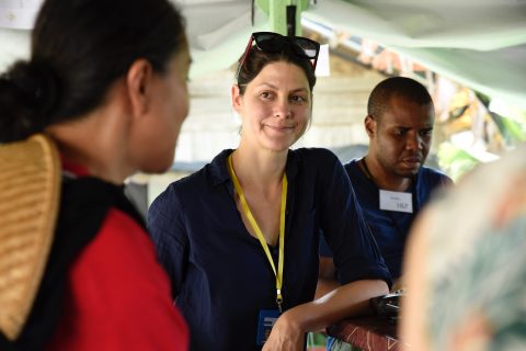 A woman in a blue shirt wearing a yellow landyard looks toward the camera. A man with a nametag is in the background.