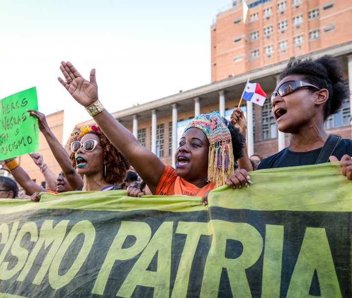 Three women hold a banner, the woman in the middle raises her arm to the sky