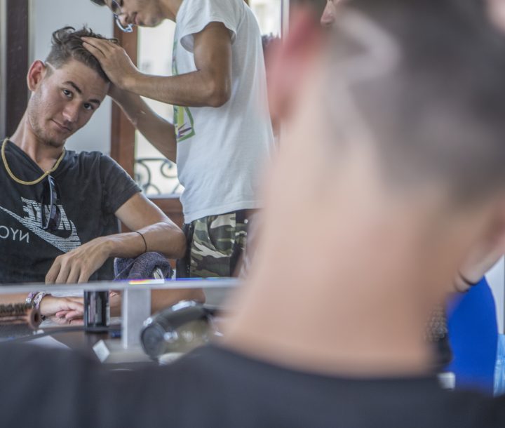 A man sits in a barber's chair looking at his reflection in the mirror, while a man cuts his hair with a razor.