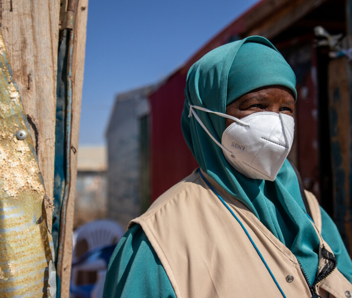 A woman wearing a face mask looks into the distance. She is wearing a turquoise uniform and head covering and a beige vest..