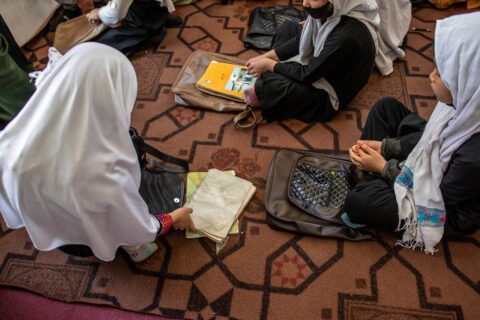 Three children sit cross legged on a decorative mat with their school books and bags at their feet.