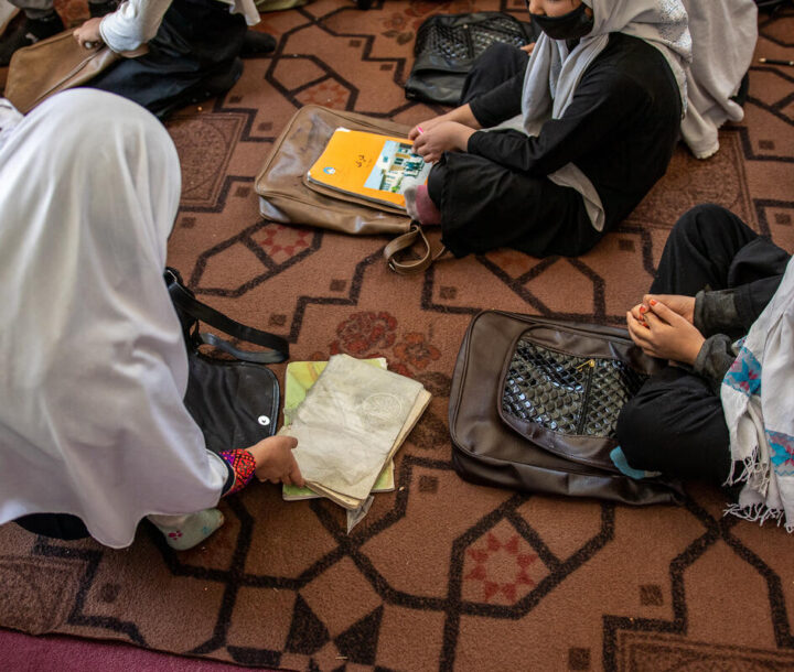 Three children sit cross legged on a decorative mat with their school books and bags at their feet.