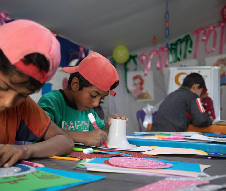 Two adolescent boys wearing red caps back to front sit at a table with textas in their hands. They look at a sheet of paper on the table.