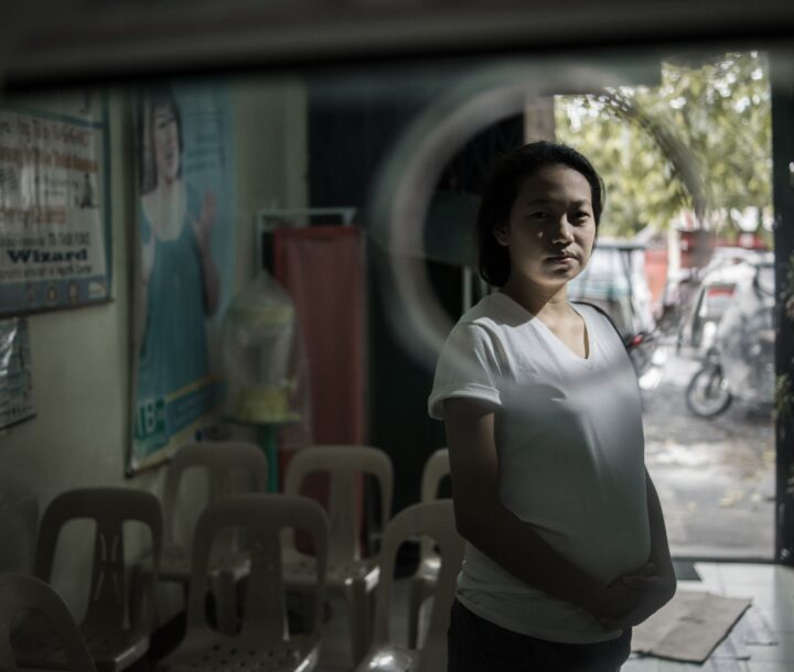 A pregnant women dressed in a white t-shirt looks through a glass window at the camera. There is a row of plastic chairs behind her.