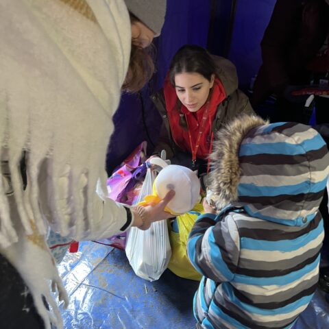 A woman wearing a jacket and red scarf is crouched down. Another woman and a child in a jacket face her and hold items from a bag.