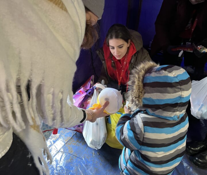 A woman wearing a jacket and red scarf is crouched down. Another woman and a child in a jacket face her and hold items from a bag.