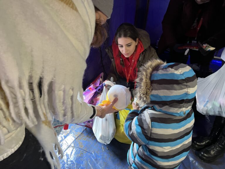 A woman wearing a jacket and red scarf is crouched down. Another woman and a child in a jacket face her and hold items from a bag.