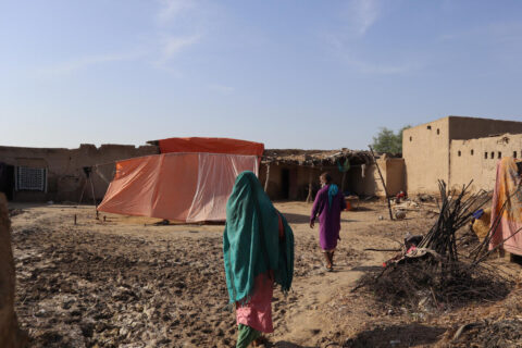 A woman wearing a green cloth walks away from the camera. There is a man and a plastic tent in the background