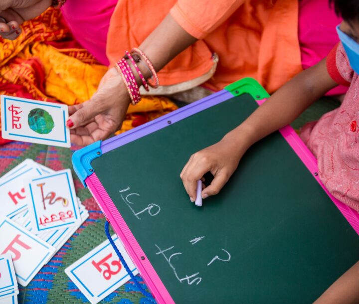 A child in a pink shirt wearing a face masks sits and writes on a chalk board. A woman next to them holds a card with a picture and writing on it.