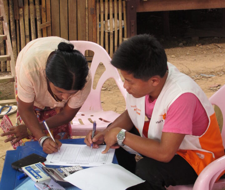 A man wearing a white vest over a pink t-shirt sits at a table. A woman leans over the table to sign a document