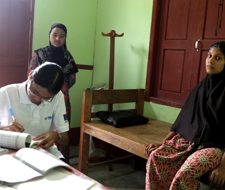 A woman in a white uniform sits at a wooden desk writing on a notepad, while another woman sits on a stretcher bed.