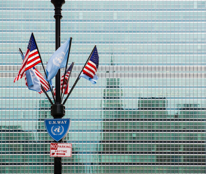 A pole with multiple American and UN flags hangind from it is positioned in front of a building with a reflective facade. The reflection of a city skyline is displayed on the building.
