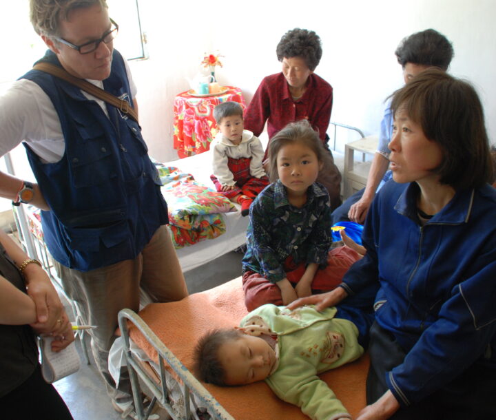 A woman sits on a hospital bed holding the hand of a small child. Another person in a blue vest faces the woman as a small child looks on