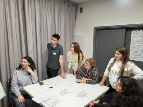 Five people gather round a square table. They are all looking off to the left hand side.