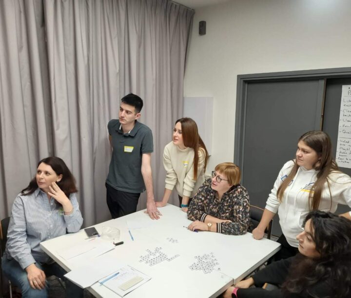 Five people gather round a square table. They are all looking off to the left hand side.