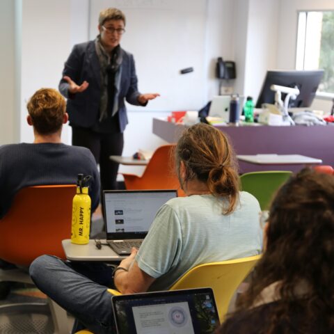 A teacher stands infront of a group of students sitting with their laptops open.