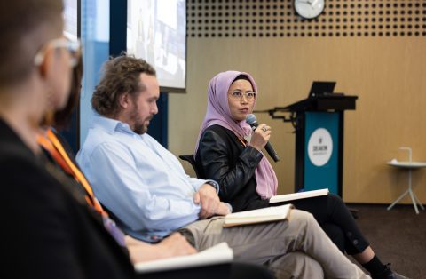 Three people sit on chairs side-on. A woman in a purple head scarf is talking into a microphone.