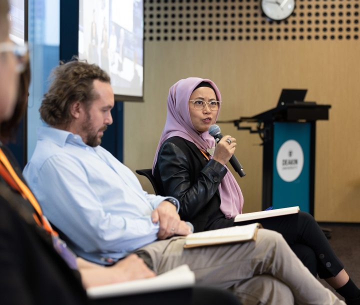 Three people sit on chairs side-on. A woman in a purple head scarf is talking into a microphone.