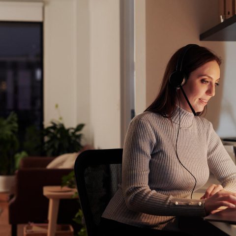 A woman in a brown turtleneck jumper sits at a desk. She is wearing headphones with an attached microphone.