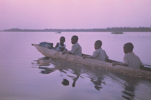 Four children sit in a canoe and float through the water. There is a pink tinge to the sky and water.