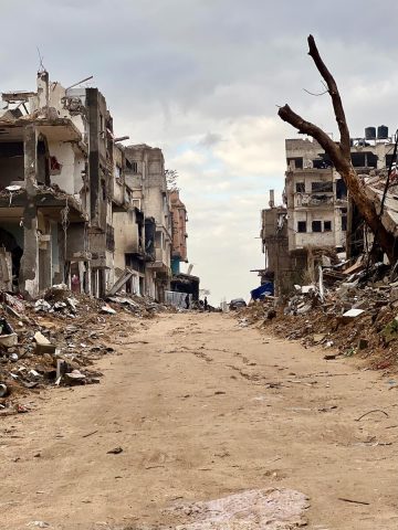 A dirt road with destroyed buildings lining either side. The sky is dark grey with clouds overhead.